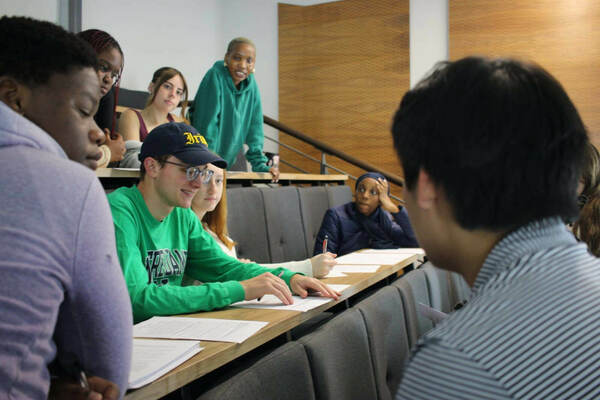 EESA Study Abroad Program. Students wearing a variety of colorful clothing sit in a lecture hall, reviewing papers and interacting with each other. One student in a green Notre Dame sweatshirt gestures towards papers on the desk.