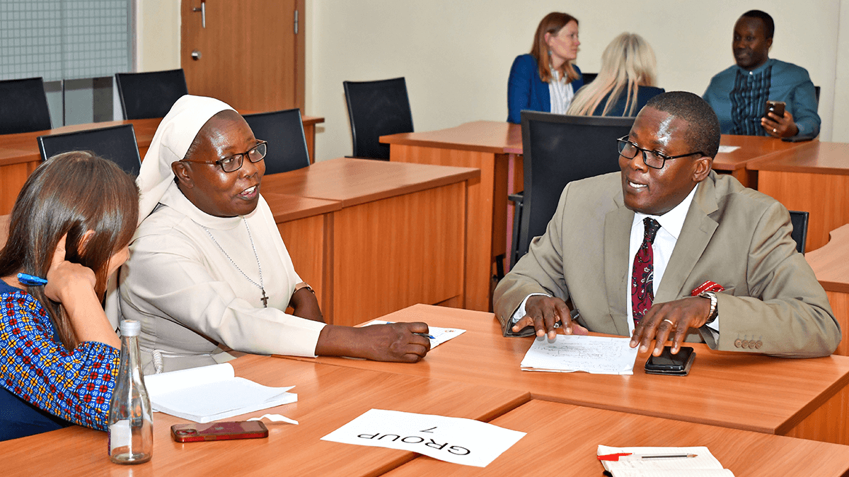 Three delegates at the Pamoja Summit. A nun wearing a white habit and glasses gestures while speaking with a man in a light brown suit and red patterned tie. They sit at a table marked "Group 1" in a classroom setting, with other individuals in the background.