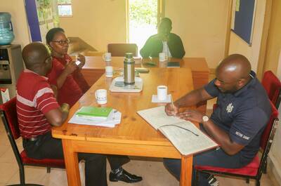 Four people sit around a wooden table. A person wearing a Notre Dame shirt writes in a ledger.  Another person gestures while speaking, and two others observe the conversation.  Mugs, a thermos, and papers are on the table.