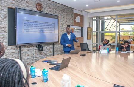 A man in a blue suit gestures while presenting on polymer technologies at Strathmore University.  A screen behind him displays the University of Notre Dame logo and a slide titled "Why Am I Here?