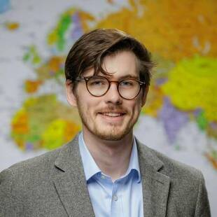 Kevin Mann. A smiling young man with light brown hair and a beard wears brown-framed glasses, a light blue shirt, and a gray blazer, standing in front of a colorful world map.